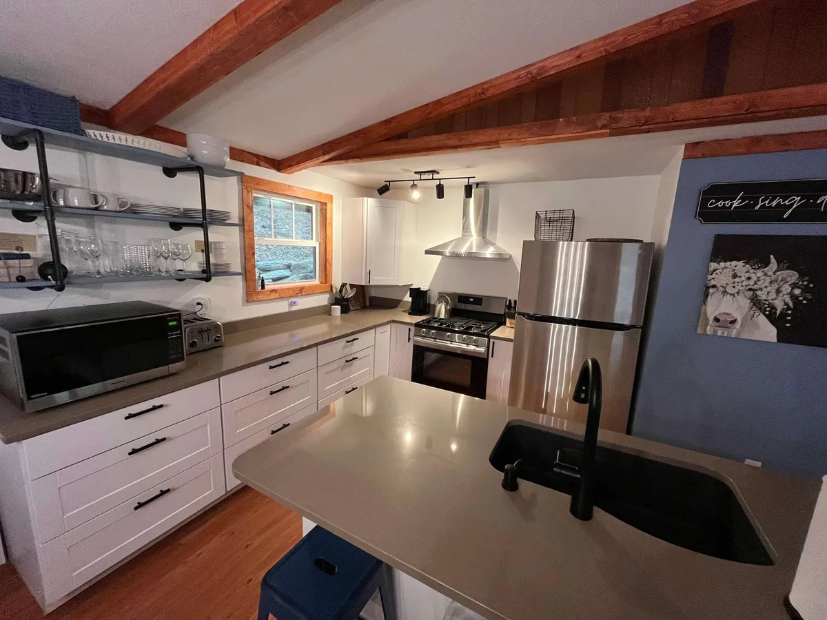 This image features a bright kitchen with a central island, stainless steel appliances, open shelving displaying glassware, and a window pouring in natural light. The scene emphasizes abundant counter space and a welcoming layout, making it perfect for cooking and casual conversation.
