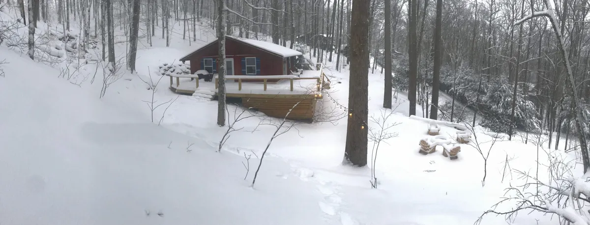 A charming cabin sits amid a snow-covered landscape, framed by trees dusted in white. The scene evokes the feeling of a secluded mountain retreat where guests can experience both adventure and quiet, peaceful moments. The mood is tranquil and inviting, emphasizing the winter wonderland aspect.