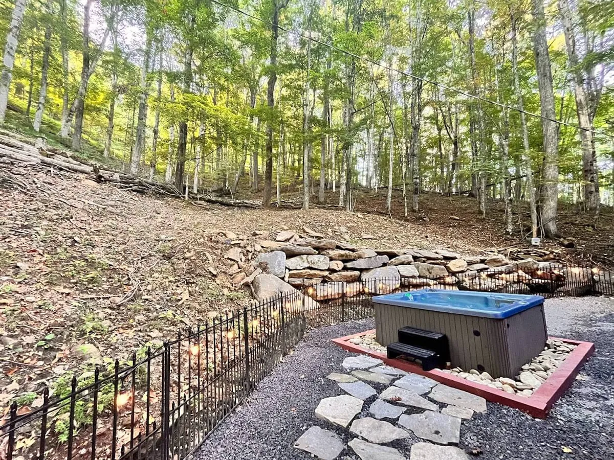A bubbling hot tub sits on the deck, surrounded by trees and a rustic rock wall. The natural setting creates a tranquil atmosphere, making it a perfect place to relax while soaking in the fresh mountain air.