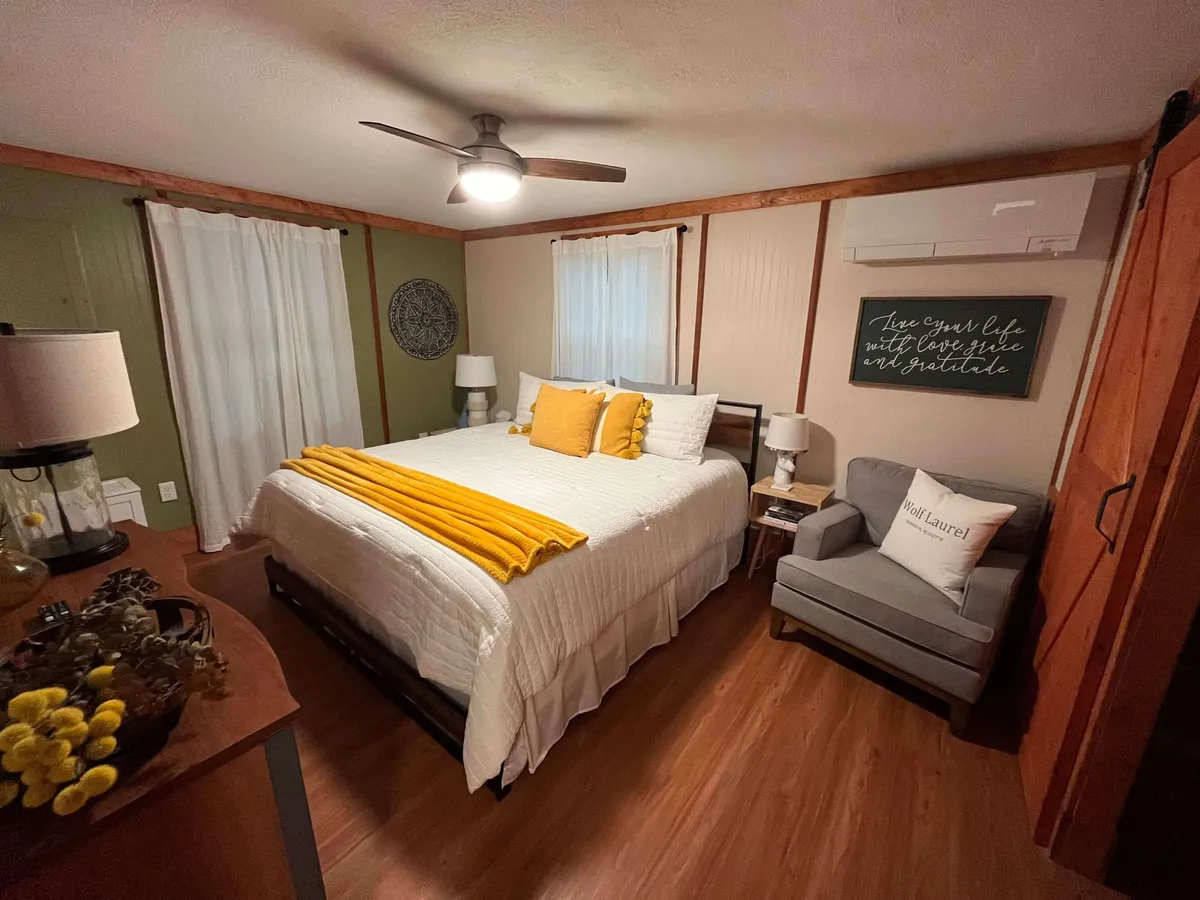 A tranquil bedroom featuring a bed with a yellow throw blanket, an accent chair with a pillow beside the window, and decorative wall art. Natural light streams in, creating a peaceful retreat vibe, while the setup suggests space for relaxing or reading quietly.