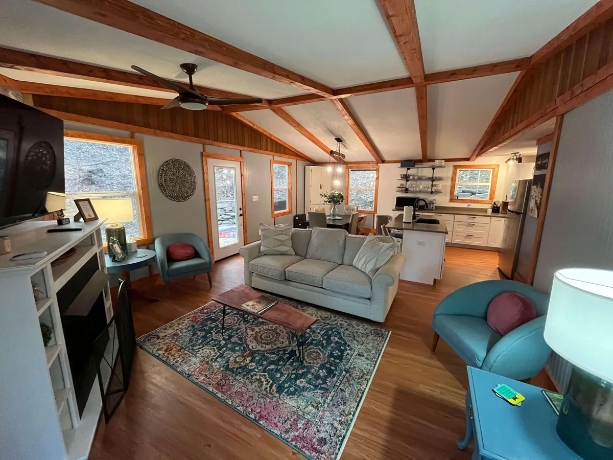 An open-plan living and dining space with a comfortable sofa, modern dining area, and a sleek kitchen in the background. Large windows allow natural light to pour in, offering glimpses of scenic outdoor views. The arrangement encourages easy interaction between living, dining, and cooking areas.