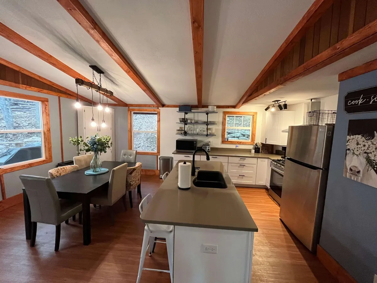 A cozy, well-lit space featuring a dining table adjacent to a kitchen island, with large windows and stylish lighting fixtures. Appliances and seating make the area ideal for both meal prep and enjoying meals together.