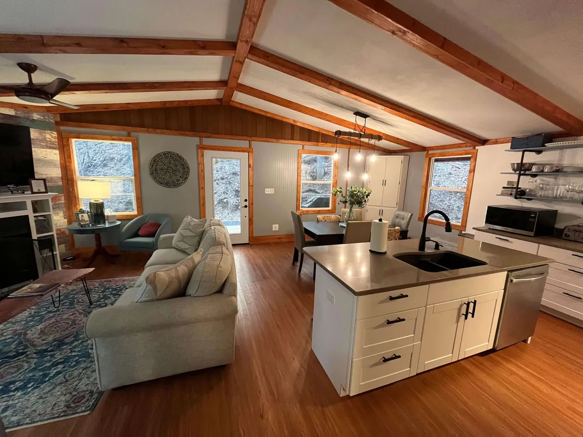 A spacious, modern interior showcasing a bright living area with a sofa, dining table, kitchen island, and a striking fireplace. Expansive windows frame beautiful mountain views, making nature an integral part of the shared space. The decor is cozy yet contemporary, perfect for both relaxation and entertaining.