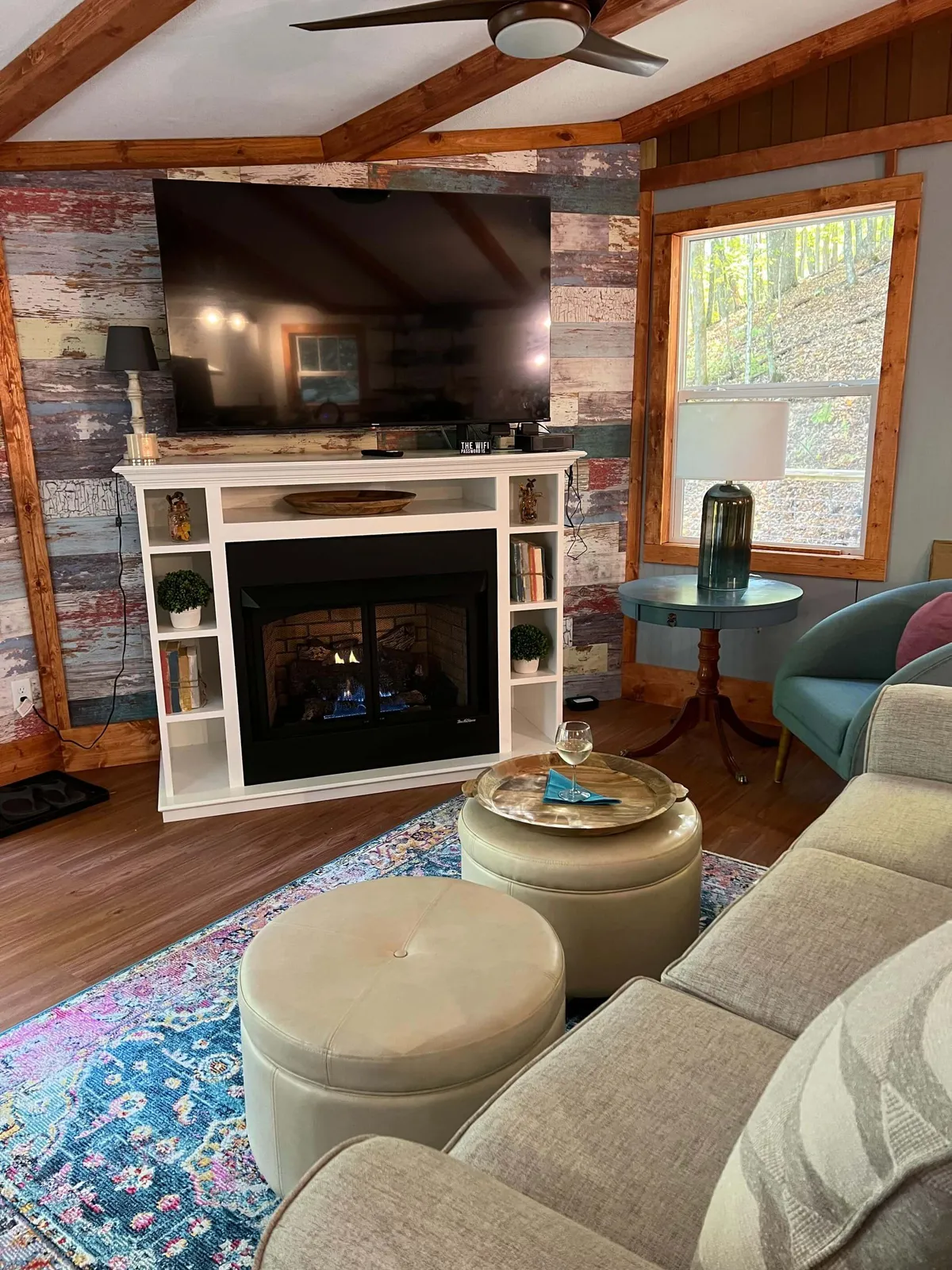 A cozy great room featuring a large stone fireplace as the centerpiece, surrounded by a plush sofa, decorative area rug, and a rustic wooden coffee table. Warm lighting and tasteful decor foster a welcoming atmosphere, while a television above the mantel and a table nearby hint at relaxing evenings and game nights.