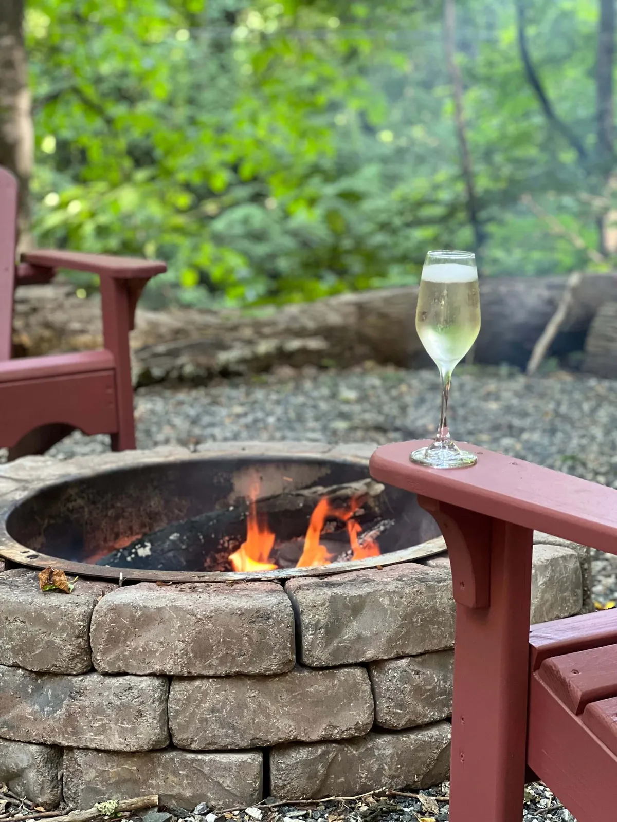An outdoor fire pit area with adirondack chairs and a sparkling drink set atop the table. The area is surrounded by trees and captures the warmth of sunset, ideal for gathering with friends or enjoying quiet evenings.