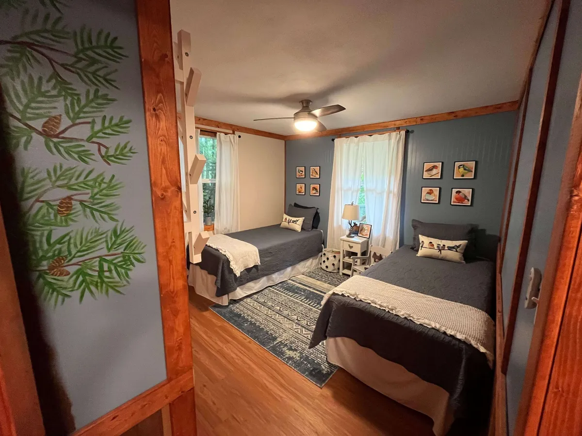 A charming bedroom with two twin beds, bird-themed wall art, a mural of pine trees, and soft lighting, creating a peaceful and cozy space. The setup suggests flexibility for families, children, or friends sharing the room.