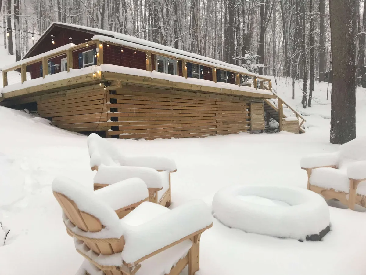 The image showcases a charming wooden cabin nestled in a snowy landscape, with inviting snow-covered chairs arranged around a glowing fire pit set against the backdrop of the Blue Ridge Mountains. The scene evokes warmth and togetherness, with the promise of stargazing, winter fun, and cozy nights spent sharing stories or hot cocoa by the fire.