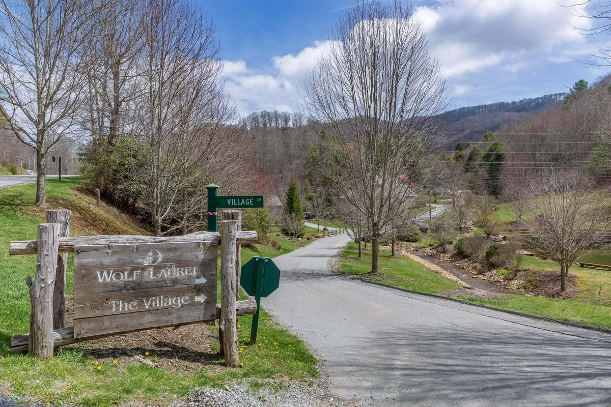 A rustic wooden sign marks the entrance to a village, with a road leading into the distance, surrounded by tall trees and mountain backdrops. The scene suggests boundless paths for hiking or biking, and a sense of arrival and discovery within the community.